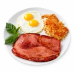 The Traditional breakfast plate featuring a thick slice of grilled ham, two sunny side up eggs, fresh basil, and golden hashbrowns at The Eggsquisite Cafe