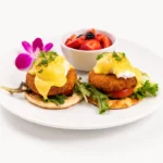 Crab Cake Benedict at Eggsquisite Cafe with poached eggs, crab cakes, spring mix, tomatoes, naan pita bread, hollandaise sauce, and chives.
